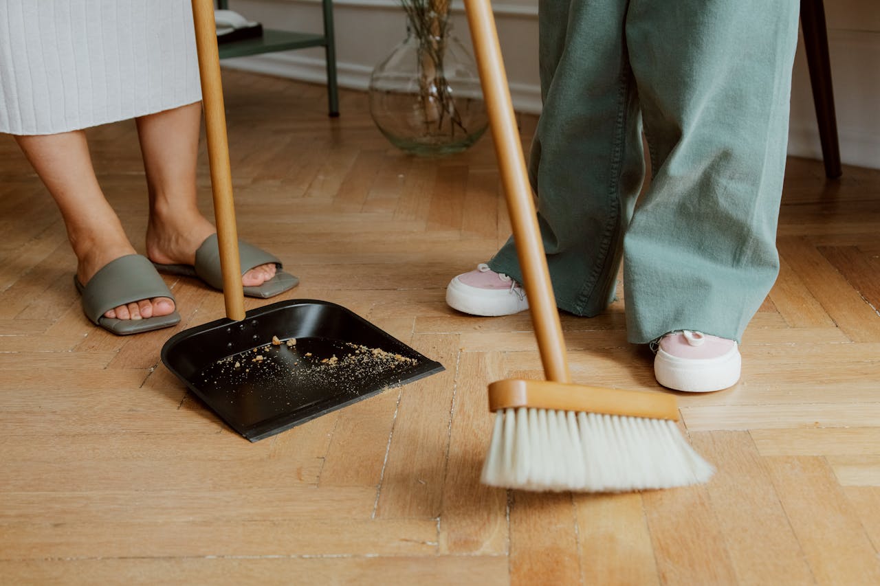 hero-img-02 Close-up of people cleaning wooden floor with broom and dustpan indoors.