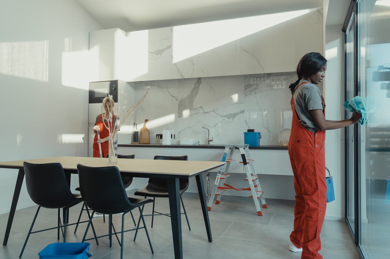 about-01 Two women in work clothes cleaning a kitchen with glass doors.